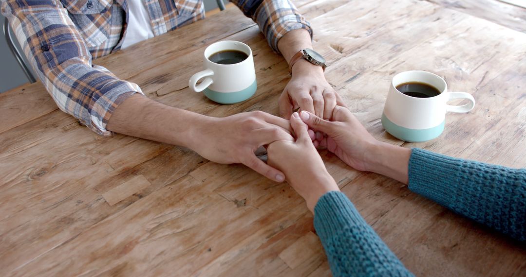 A close-up of two partners holding hands across a wooden coffee table with two mugs, suggesting a quiet reconciliation moment