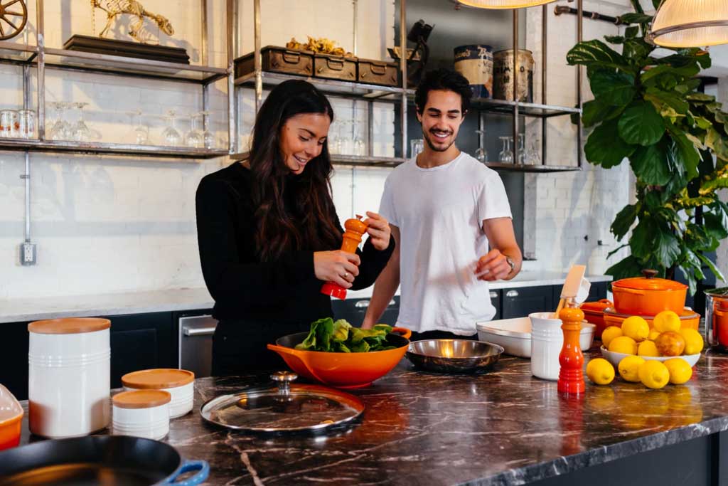 A couple cooking together in a home kitchen in the evening, one person chopping vegetables while the other stirs a pan under warm overhead light