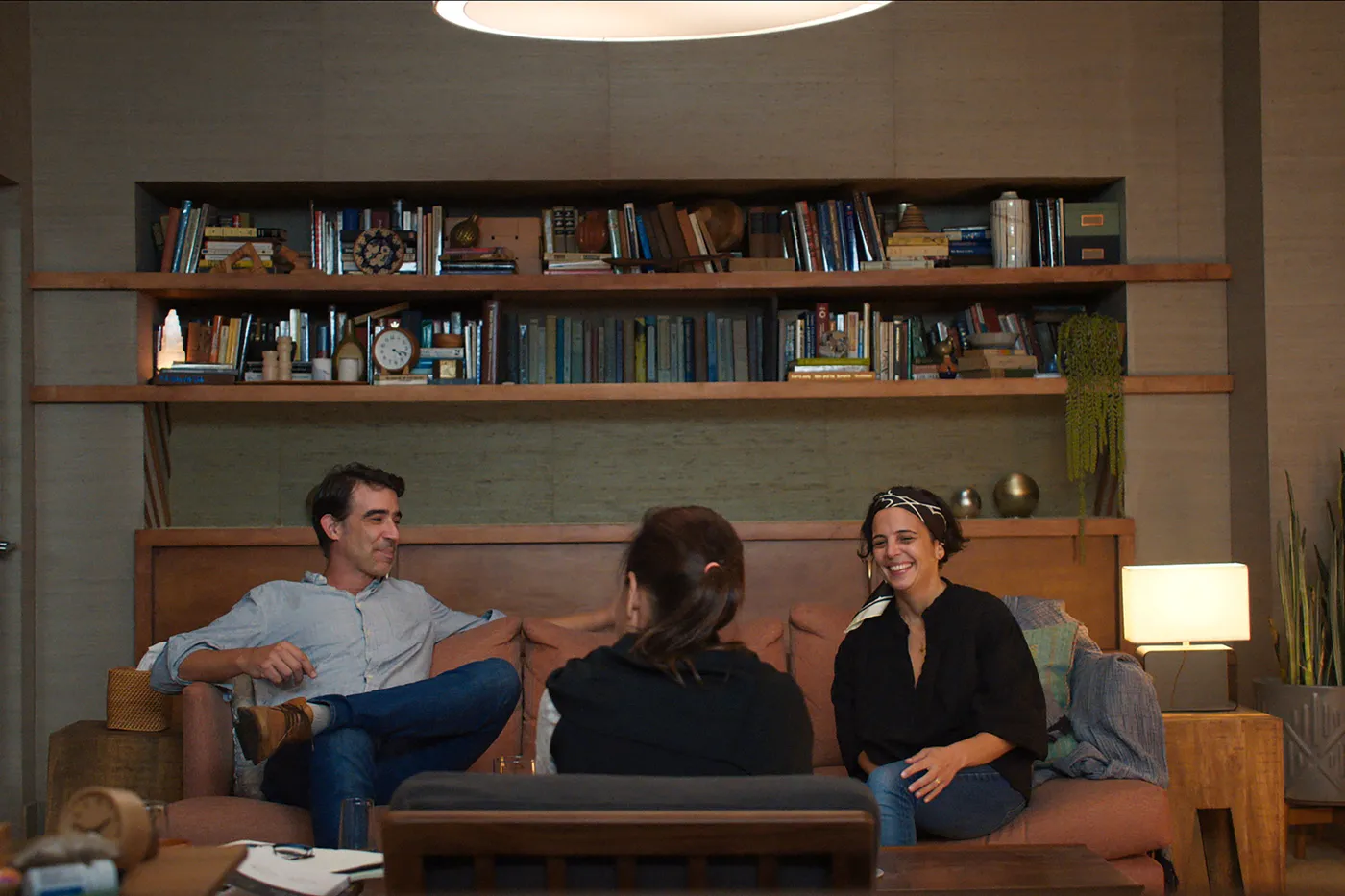 A couple seated on a sofa in a calm therapy office, speaking with a therapist in an armchair, natural light through a window