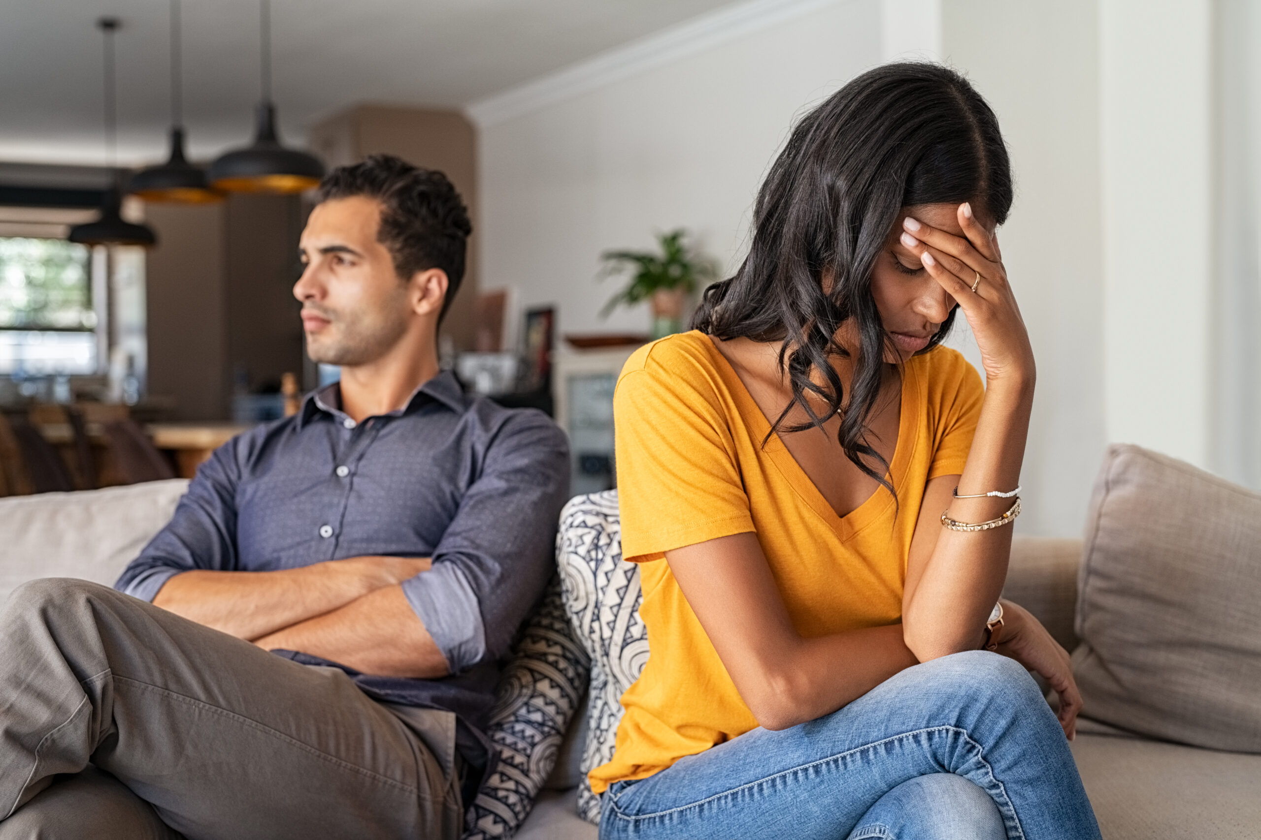 A couple sitting back to back on a living room couch, both looking away with crossed arms, natural indoor lighting