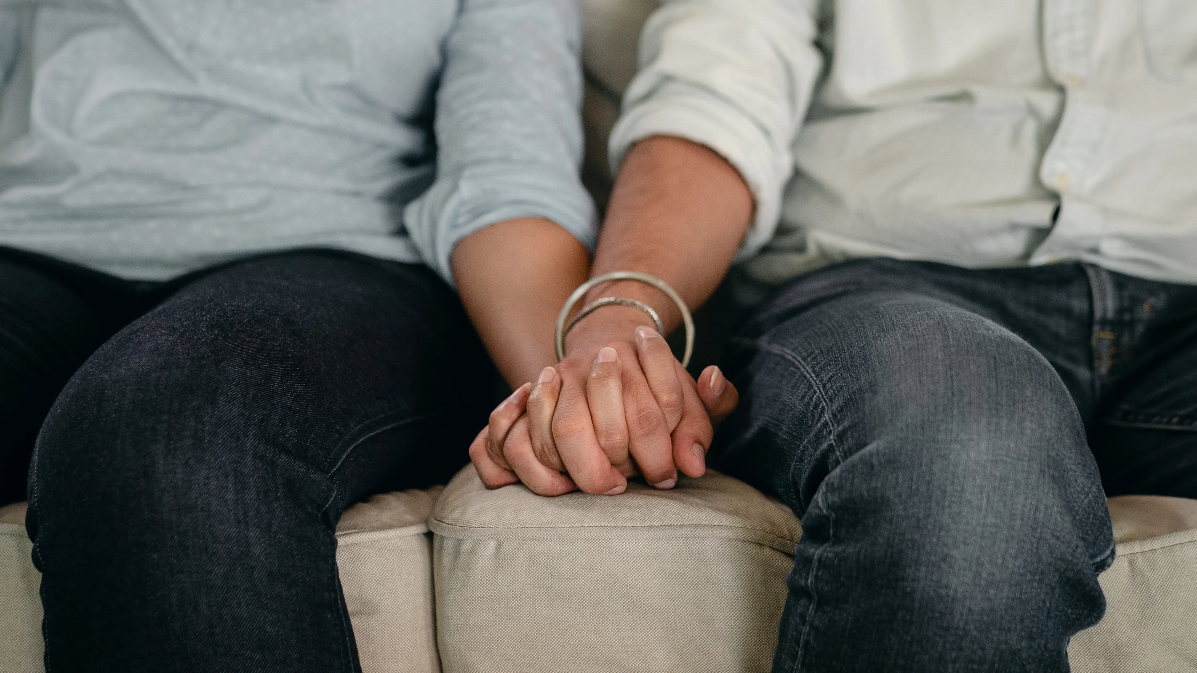 A couple sitting close together on a living room sofa, one partner gently holding the other’s hand while listening, natural light coming through a window