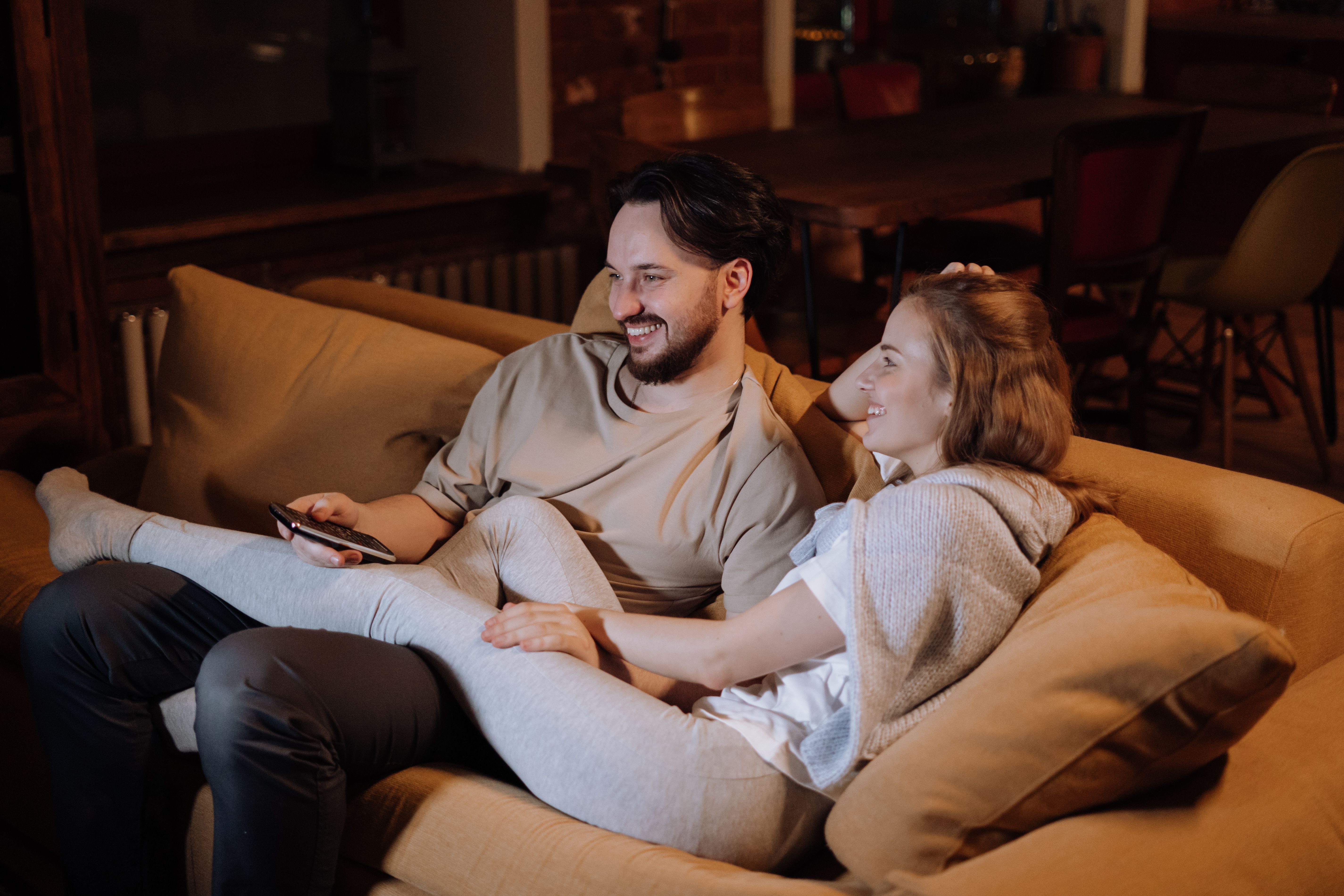 A couple sitting on opposite ends of a living room couch in the evening, looking tired and quiet with a TV glow in the room