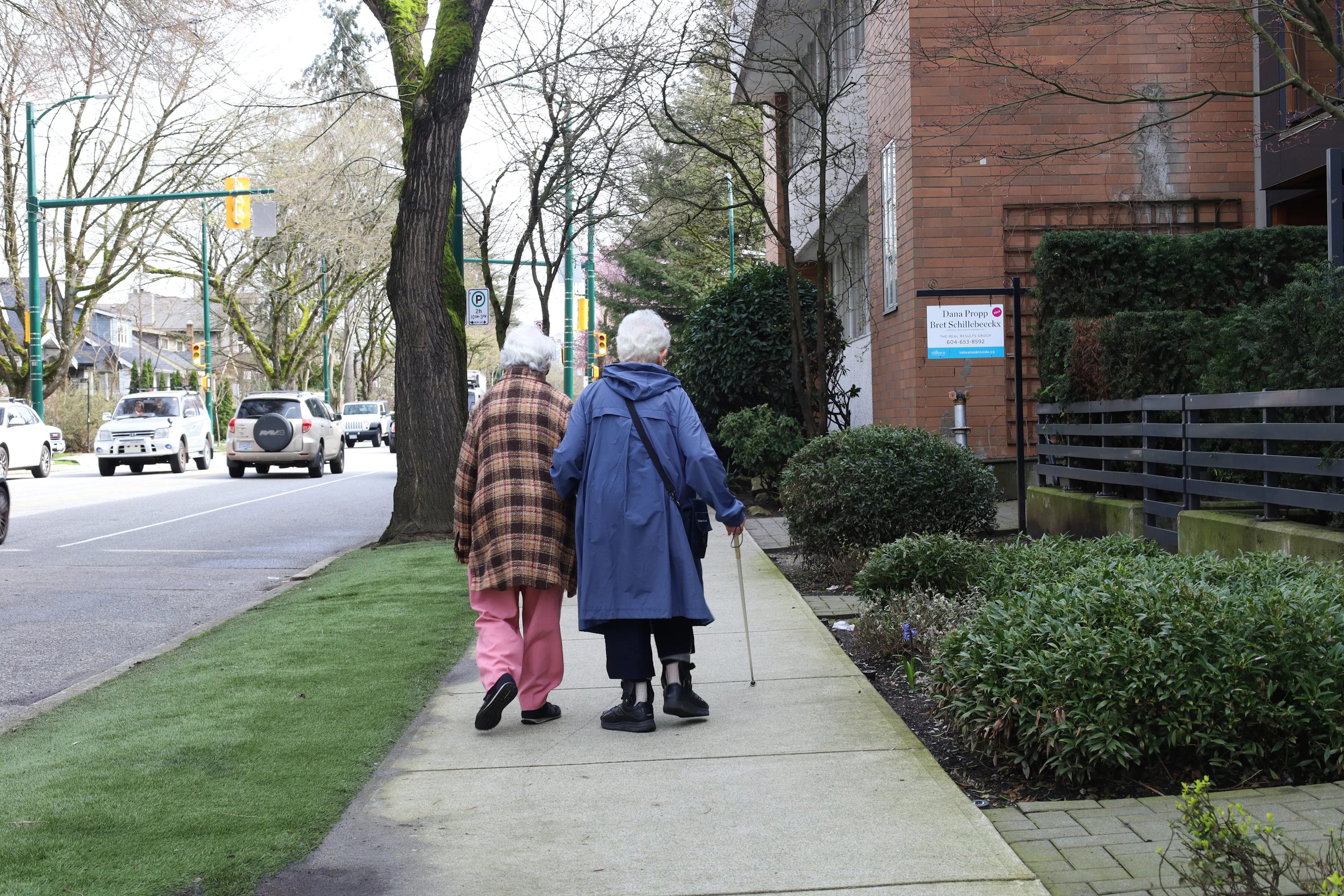 A couple walking side by side on a quiet residential sidewalk in the early evening, wearing casual clothes, hands lightly brushing as they walk