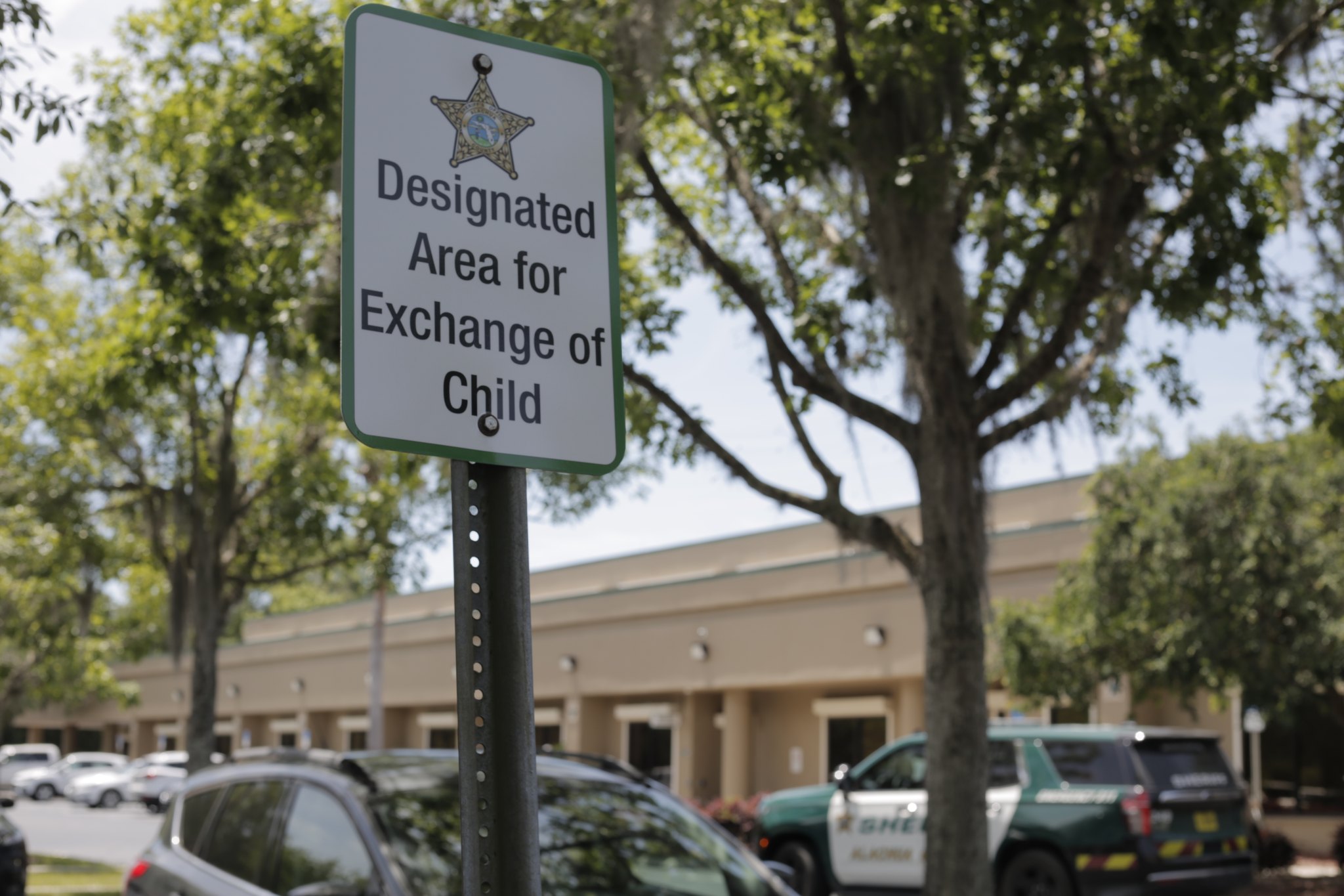 A divorced mother and father doing a calm child handoff in a public park parking area during daytime, with their child holding a backpack between them