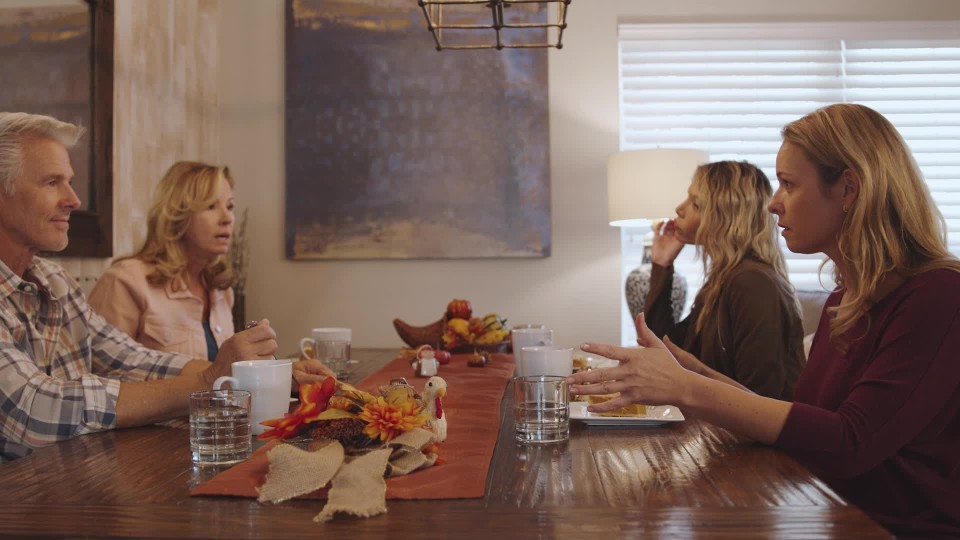 A family sitting around a kitchen table in visible tension, with one parent looking frustrated and a teenager looking away, candid home photograph