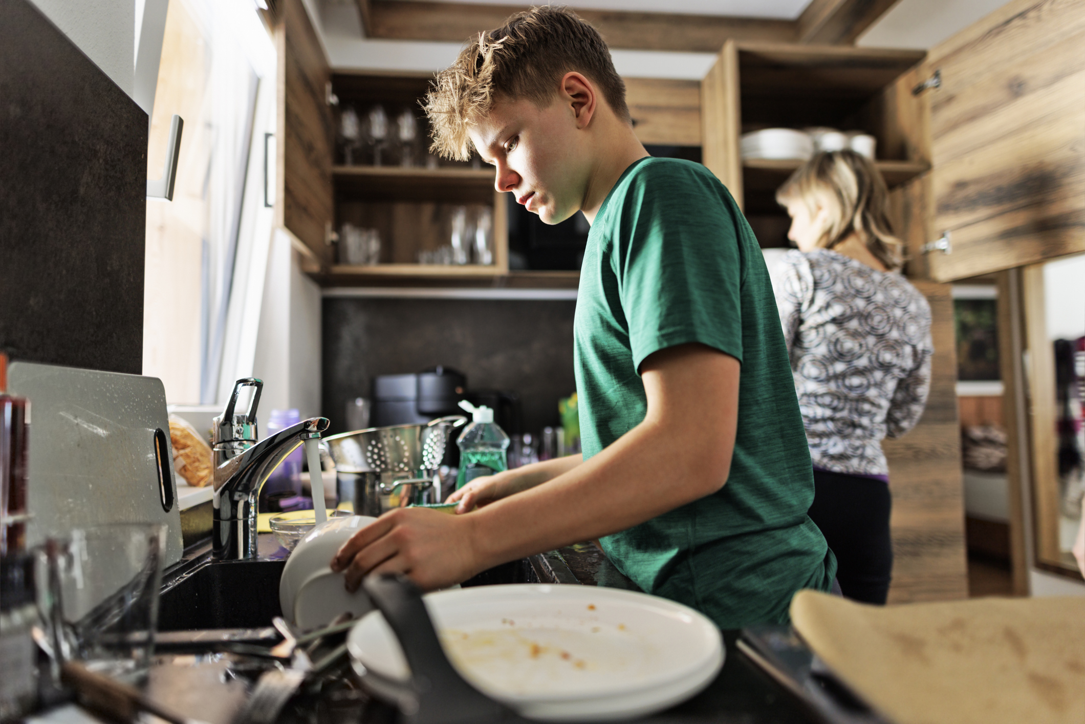 A parent and a teenager dry dishes together at a kitchen counter, smiling in a relaxed everyday moment.