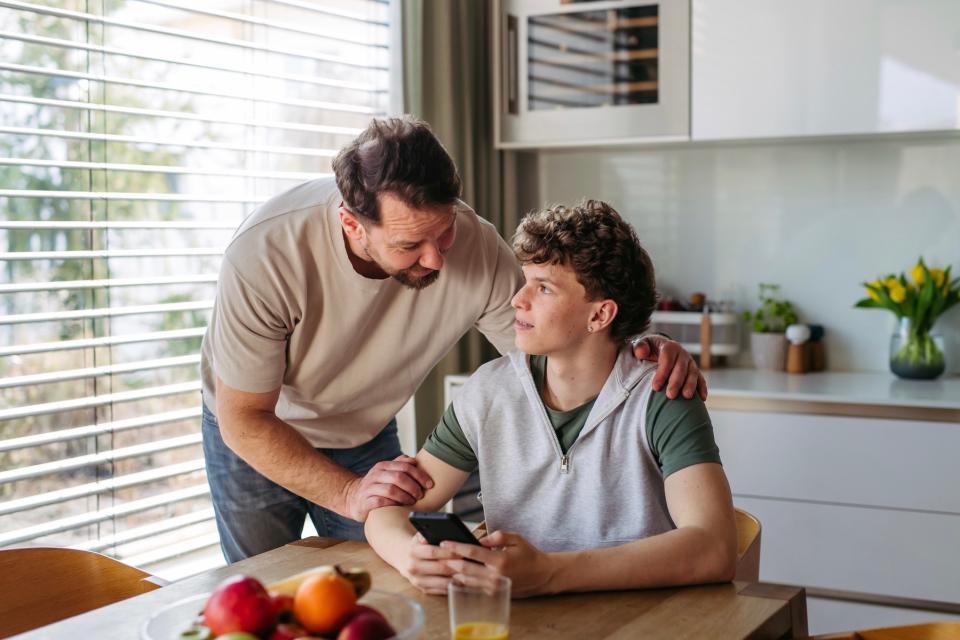 A parent and a teenager sitting at a kitchen table talking, with notebooks and a glass of water on the table