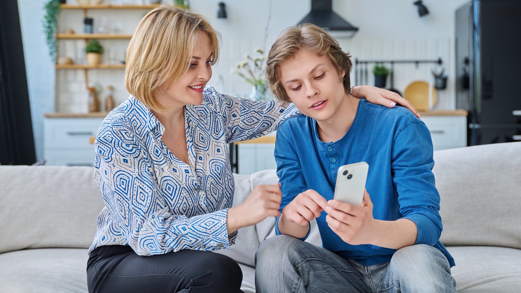 A parent and a teenager sitting on a couch looking at a smartphone together during a calm conversation