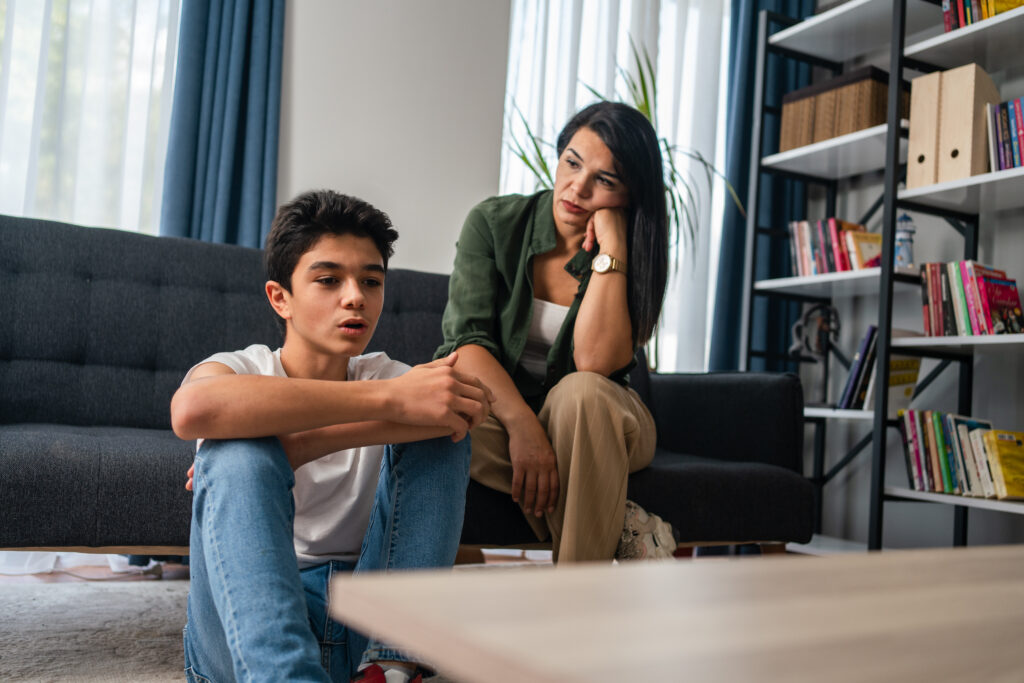 A parent and a teenager sitting on a living room couch in soft evening light, facing each other with calm attentive expressions