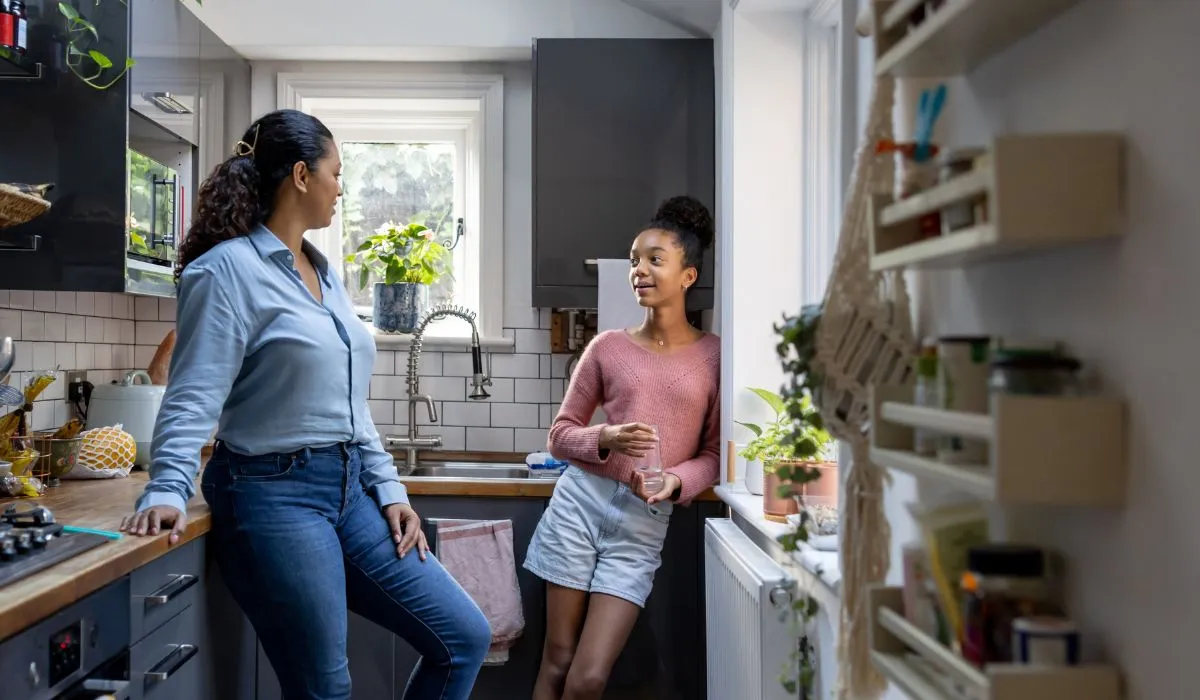 A parent and a teenager stand side by side in a kitchen, talking casually while the teen snacks.