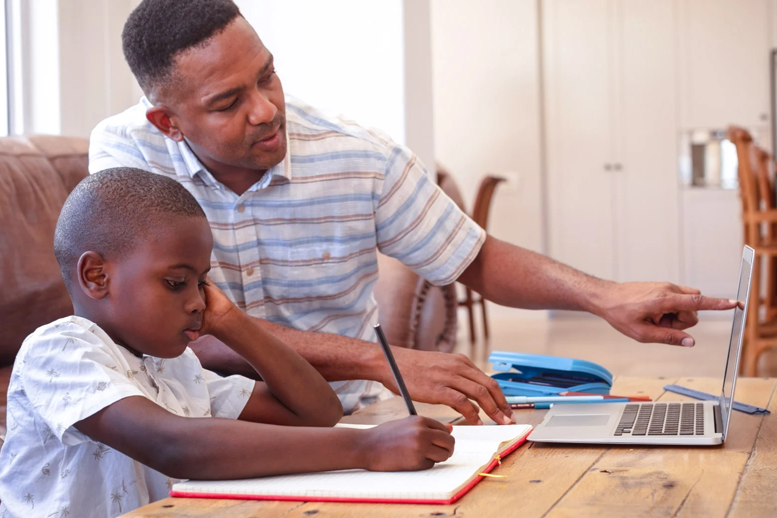 A parent sitting with a middle school-aged child at a kitchen table, calmly looking over homework papers with notebooks and pencils spread out