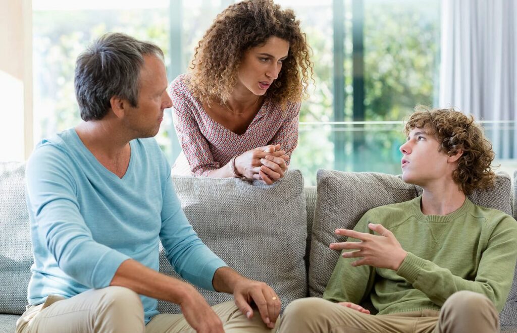 A parent sitting with a teenager on a living room sofa, both leaning slightly toward each other in a serious, supportive conversation
