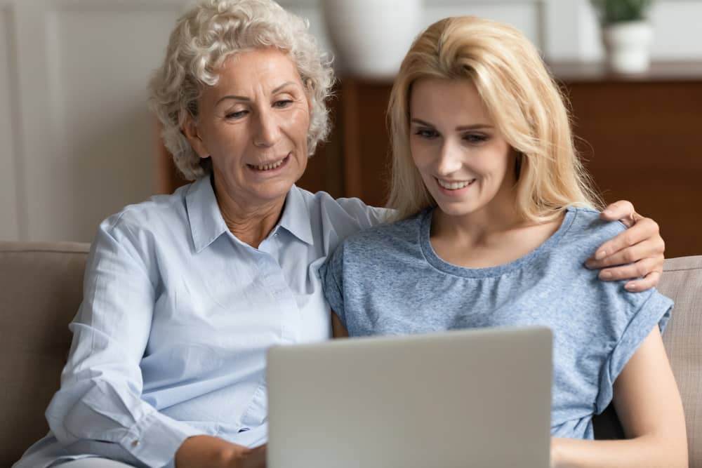 An adult daughter and her older mother sitting at a kitchen table with tea mugs, talking calmly in soft afternoon light