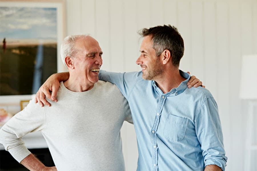 An adult son standing near a front porch, speaking calmly with his older father during a tense but respectful moment outside a suburban home