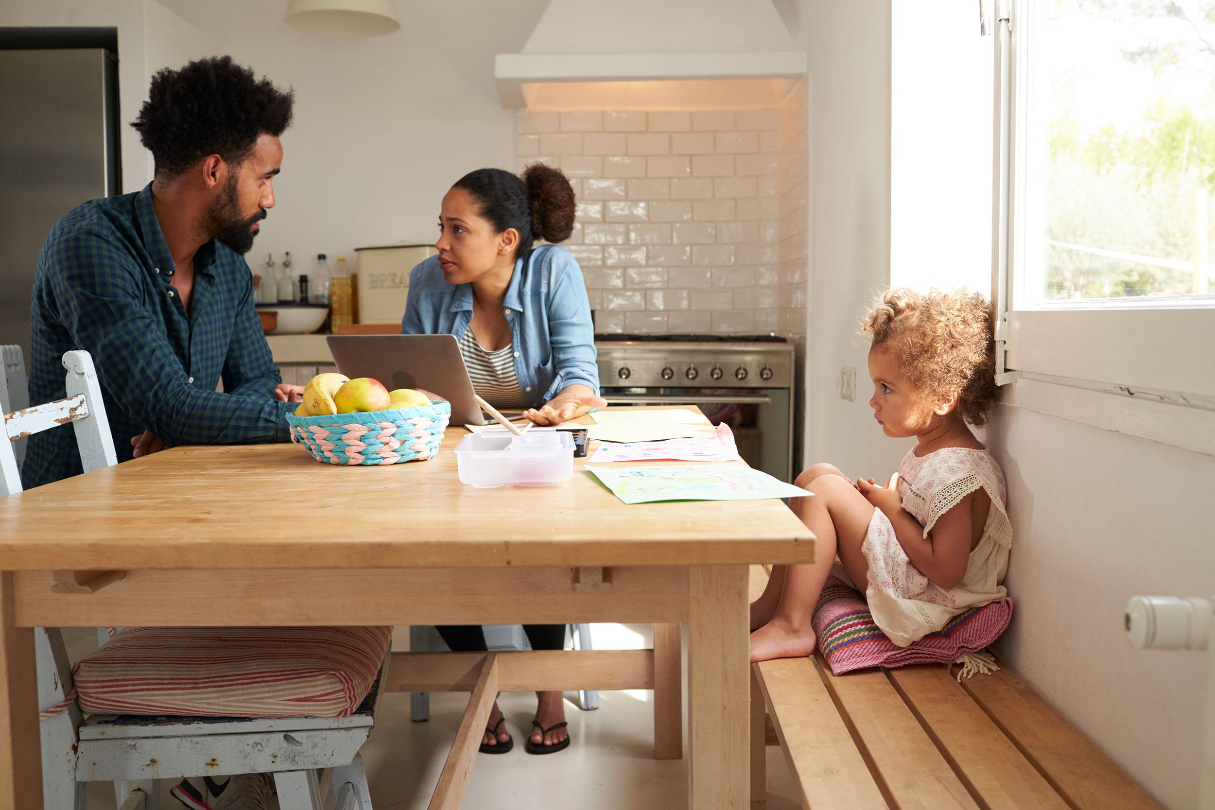 Two separated parents speaking calmly at a kitchen table while their child plays quietly in the next room